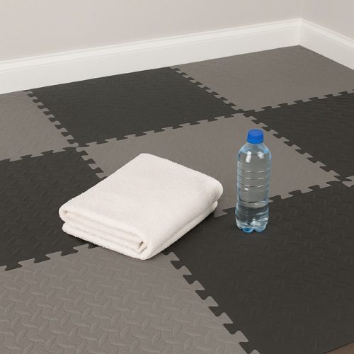 Home gym corner with grey and black EVA foam interlocking tiles in a checkerboard pattern, featuring a folded white towel and a water bottle placed on the textured floor.