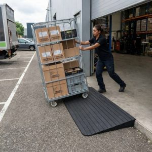 Warehouse employee pushing a loaded roll cage over a heavy-duty rubber threshold ramp at a warehouse entrance to move goods safely over a raised doorstep.